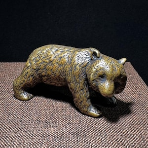 Op de afbeelding: Een bronzen beerfiguur met gedetailleerde vachttextuur. De beer wordt afgebeeld in een loopstand, met zijn hoofd iets naar beneden. De sculptuur staat tegen een donkere achtergrond, waardoor de metallic afwerking van de beer wordt benadrukt.