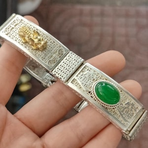 May include: Silver bracelet with intricate filigree design. Features a gold-colored figurine and a large oval green gemstone. The bracelet is held in a hand, showcasing its detailed craftsmanship and elegant style.