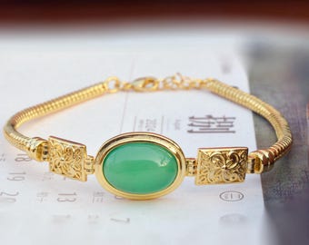 May include: Gold-toned bracelet featuring a central oval-shaped green stone. The bracelet has a textured chain design with decorative square accents on either side of the stone. The clasp is also gold-toned.