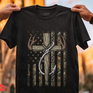 May include: Black t-shirt featuring a graphic design with a cross, antlers, and a fishing hook, set against a camouflage and star-patterned American flag. The design incorporates brown, beige, and white elements.