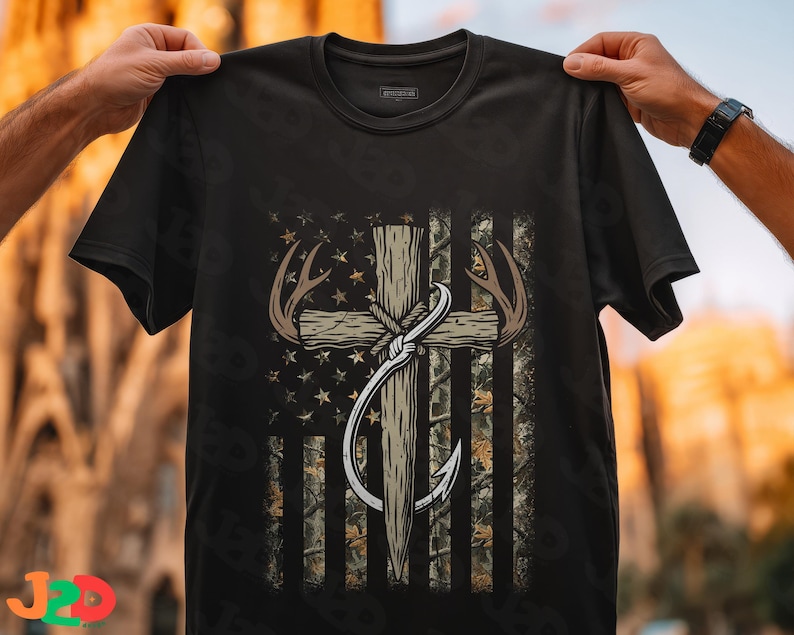 May include: Black t-shirt featuring a graphic design with a cross, antlers, and a fishing hook, set against a camouflage and star-patterned American flag. The design incorporates brown, beige, and white elements.