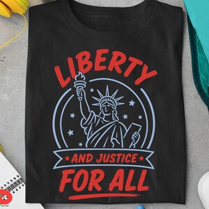 May include: A black t-shirt featuring a graphic of the Statue of Liberty in light blue, surrounded by stars. The text in red reads LIBERTY at the top and AND JUSTICE FOR ALL on a banner below the statue, with a circular border surrounding the design.
