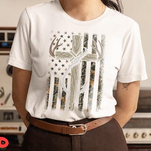 May include: White t-shirt featuring a graphic design with a cross, antlers, and a fishing hook, set against a backdrop resembling the American flag. The design incorporates camouflage and stars.