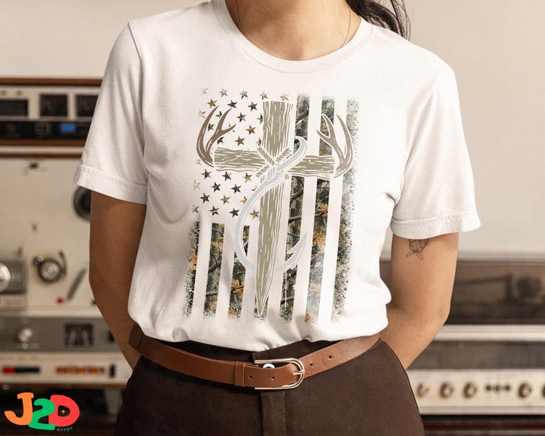 May include: White t-shirt featuring a graphic design with a cross, antlers, and a fishing hook, set against a backdrop resembling the American flag. The design incorporates camouflage and stars.