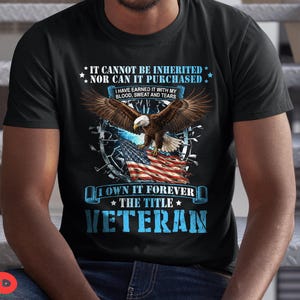 May include: Black t-shirt with a graphic featuring an eagle with outstretched wings above an American flag. Text includes "IT CANNOT BE INHERITED NOR CAN IT PURCHASED" and "I OWN IT FOREVER THE TITLE VETERAN."