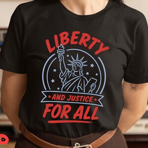 May include: A black short-sleeved t-shirt featuring a graphic of the Statue of Liberty in light blue, surrounded by stars. The text LIBERTY is printed in red above, and AND JUSTICE FOR ALL is printed in red below the statue on a light blue banner.