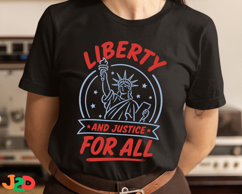 May include: A black short-sleeved t-shirt featuring a graphic of the Statue of Liberty in light blue, surrounded by stars. The text LIBERTY is printed in red above, and AND JUSTICE FOR ALL is printed in red below the statue on a light blue banner.