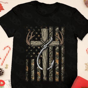 May include: Black t-shirt featuring a graphic design of a cross with deer antlers and a fishing hook, set against a camouflage American flag background. The design incorporates stars and stripes, creating a patriotic and outdoorsy theme.