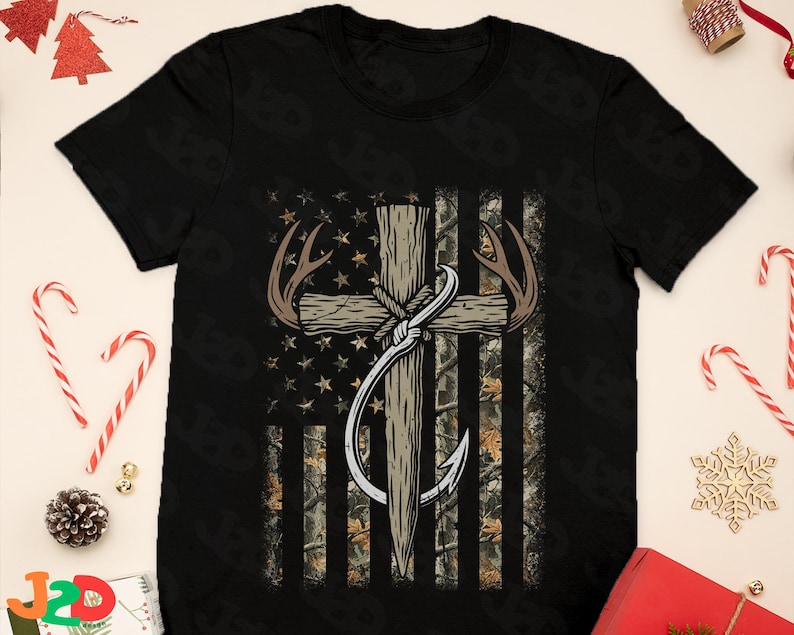 May include: Black t-shirt featuring a graphic design of a cross with deer antlers and a fishing hook, set against a camouflage American flag background. The design incorporates stars and stripes, creating a patriotic and outdoorsy theme.