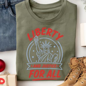 May include: An olive green t-shirt featuring a graphic of the Statue of Liberty in light blue, surrounded by stars. The text "LIBERTY" appears in red above the graphic, and "AND JUSTICE FOR ALL" is printed in red below the statue illustration.