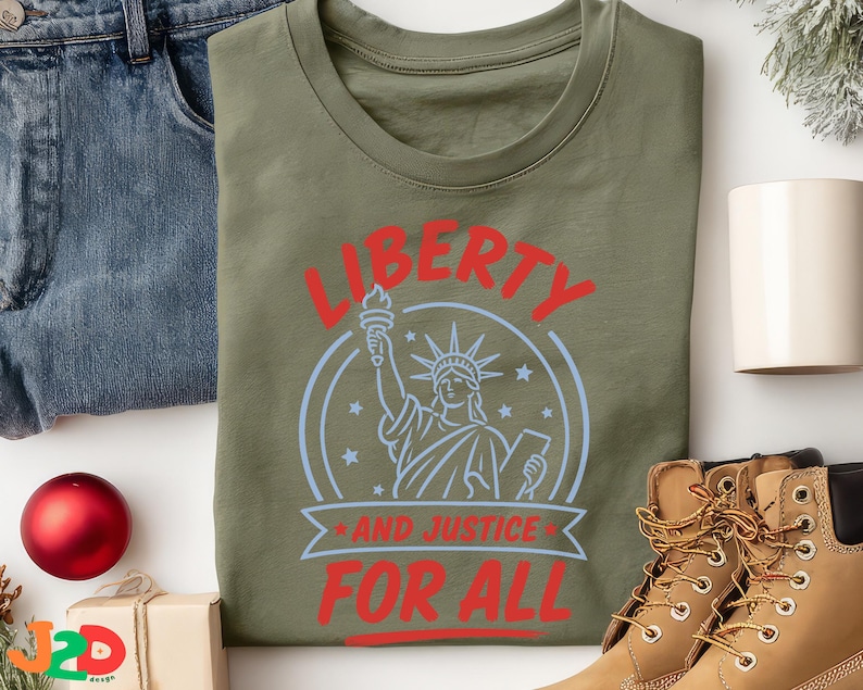 May include: An olive green t-shirt featuring a graphic of the Statue of Liberty in light blue, surrounded by stars. The text "LIBERTY" appears in red above the graphic, and "AND JUSTICE FOR ALL" is printed in red below the statue illustration.