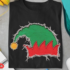 Op de afbeelding: Een zwart T-shirt met korte mouwen voorzien van een afbeelding van een groene elfenmuts met een rode, gekartelde rand en een gele pompon. Het ontwerp heeft een gerafelde, versleten textuur met zichtbare witte draden langs de randen.