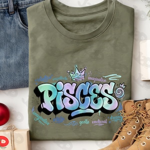 May include: An olive green t-shirt featuring the word "PISCES" in a graffiti-style design with blue and purple hues. The design includes a crown and the words "imaginative", "artistic", "gentle", and "emotional".