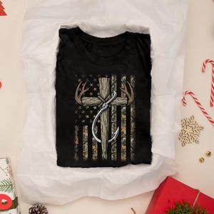 May include: Black t-shirt featuring a graphic of an American flag with a cross, antlers, and a fishing hook. The flag design incorporates camouflage and stars. The shirt is folded on white tissue paper, surrounded by Christmas decorations.