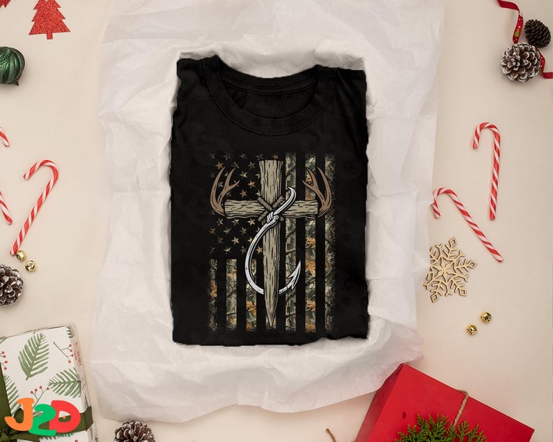 May include: Black t-shirt featuring a graphic of an American flag with a cross, antlers, and a fishing hook. The flag design incorporates camouflage and stars. The shirt is folded on white tissue paper, surrounded by Christmas decorations.