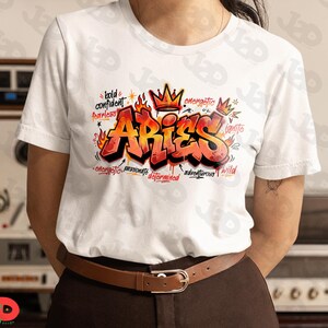 May include: White t-shirt featuring a graffiti-style design with the word "ARIES" in red and orange, accented with flames and a crown. Additional words include "bold," "confident," "fearless," "energetic," and "wild."