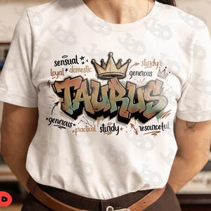 May include: White t-shirt featuring the word "TAURUS" in a graffiti style with a crown above it. The design includes words like "sensual", "loyal", "generous", and "resourceful".