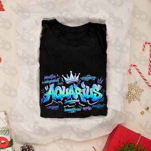 May include: Black t-shirt with a vibrant, graffiti-style design featuring the word "Aquarius" in blue and teal. The design includes a crown and words like "inventive" and "visionary". The shirt is folded on white paper, surrounded by holiday decorations.