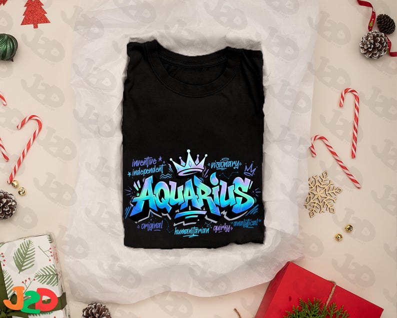 May include: Black t-shirt with a vibrant, graffiti-style design featuring the word "Aquarius" in blue and teal. The design includes a crown and words like "inventive" and "visionary". The shirt is folded on white paper, surrounded by holiday decorations.