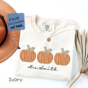 May include: An ivory t-shirt featuring a graphic of three orange striped pumpkins with green gingham stems. The text "Mrs. Smith" is printed in black cursive below the pumpkins. A blue square overlay states "FAUX PATCHWORK NOT REAL".