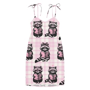 以下が含まれることがあります： ピンクと白のチェック柄のノースリーブドレス。メガネをかけて本を読むアライグマのイラストが繰り返されています。ドレスは肩で結ぶ細いストラップが付いています。