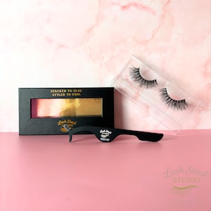 May include: A set of false eyelashes with a black applicator tool and a black box. The box has gold and pink accents and the text "STACKED TO SLAY. STYLED TO STAY." The background is pink and marble.