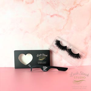 May include: A black box with a heart-shaped cutout, false eyelashes in a clear case, and a black eyelash applicator. The box and applicator have the text "Lash Stack Studio" and "Stacked to Slay, Styled to Stay."