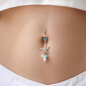 May include: A silver belly button ring featuring a starfish, pearl, turquoise bead, and seashell charm. The top gem is light blue. The jewellery is suspended from a belly button piercing.