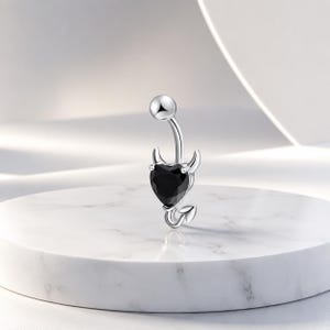 Può includere: Un piercing all'ombelico color argento con una gemma nera a forma di cuore. L'anello presenta piccole corna e un design a coda, che gli conferisce un aspetto diabolico. La sfera superiore è anch'essa argentata. Il gioiello è esposto su una superficie di marmo bianco.
