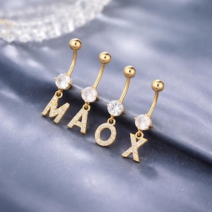 Pode incluir: Quatro piercings dourados para o umbigo que soletram a palavra "MAOX". Cada um tem um design de barra curva com uma gema redonda e transparente no centro e uma letra pendurada incrustada com pequenas pedras transparentes. O fundo é uma superfície texturizada azul-cinza.