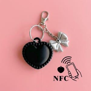 May include: A black heart-shaped keychain with a beaded border, attached to a silver-toned metal lobster clasp and a small silver bow charm. A graphic icon shows a heart and a smartphone with signal waves above the text NFC, indicating connectivity.