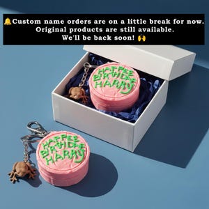 May include: Pink round keychain with "Happee Birthday Harry" in green frosting-style lettering, and a frog charm. One keychain is in a white box with blue tissue paper. The text in the image says: "Custom name orders are on a little break for now. Original products are still available. We'll be back soon!"