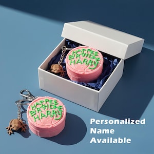 May include: Pink round keychain with green text that reads "Happy Birthday Harry". The keychain has a small frog charm. The keychain is in a white box with blue tissue paper. The text "Personalized Name Available" is on the right side of the image.