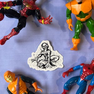 May include: A collection of Spider-Man action figures and a sticker on a light purple surface. The figures include Spider-Man in red and black, Doctor Octopus in orange and green, and the Hobgoblin. The sticker features a black and white sketch of Spider-Man.