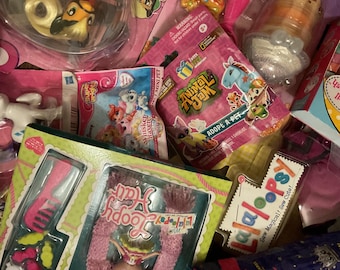 May include: A selection of children's toys, including dolls and blind bags, in various packages. The image shows toys with text such as "Animal Jam" and "Lalaloopsy". The toys are in a variety of colours, including pink, green, and blue.