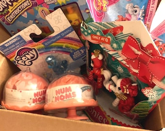 May include: A cardboard box containing a selection of children's toys, such as Shopkins, My Little Pony, and Num Noms. The toys are in their original packaging, with vibrant colours and graphics. Some packages have text like "Rainbow Dash" and "Buildable Figures!"