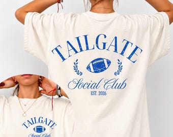 May include: An ivory t-shirt featuring blue graphics. The back displays the text TAILGATE SOCIAL CLUB EST. 2026 with a football and laurel leaves. A smaller version of the same graphic is printed on the front left chest area of the shirt.