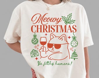 May include: An ivory t-shirt featuring a red and green graphic print. The design includes a cat wearing sunglasses, holding a wine glass, with the text "Meowy CHRISTMAS" and "Ya filthy humans!" along with holiday icons like holly and a tree.