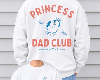 May include: A white sweatshirt featuring a back graphic with the text PRINCESS DAD CLUB in coral, a blue unicorn illustration, and the text Living in glitter & chaos. The front features the text DAD in coral with a blue crown and est. 2026.