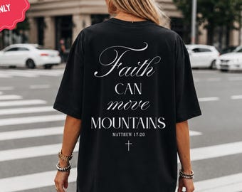 May include: A black t-shirt featuring white text on the back that reads Faith CAN move MOUNTAINS MATTHEW 17:20 with a small cross symbol below the text. The shirt has short sleeves and a loose fit, and a pink badge states BACK PRINT ONLY.