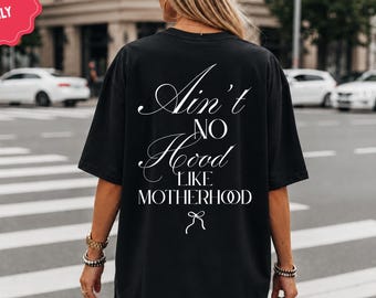 May include: A black short-sleeve t-shirt featuring white text on the back that reads "Ain't NO Hood LIKE MOTHERHOOD" with a small bow graphic underneath. The shirt is worn with a black baseball cap.