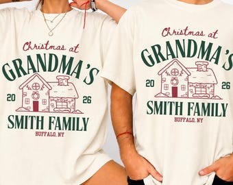 May include: An ivory short-sleeve t-shirt featuring a graphic print with the text "Christmas at GRANDMA'S", "SMITH FAMILY", "BUFFALO, NY", and the numbers "20" and "26" flanking a line-art illustration of a house decorated for the holidays.