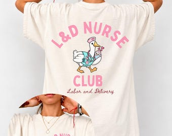 May include: An ivory t-shirt featuring a graphic of a goose dressed as a nurse holding a baby. The back displays the text L&D NURSE CLUB and Labor and Delivery in pink and brown. The front chest area features the text L&D NURSE CLUB est. 2026.
