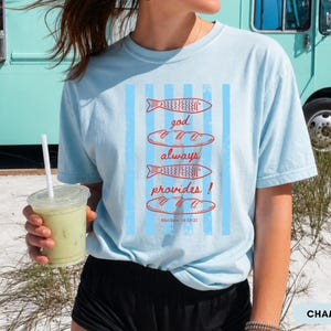 Woman wearing oversized Comfort Colors Christians shirt featuring God Always Provides graphic with fish illustrations and Matthew 14:13-21 Bible verse in vintage aesthetic style.