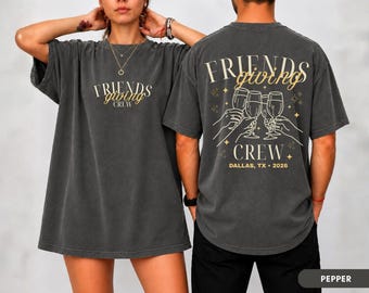 May include: A charcoal gray short-sleeve t-shirt featuring the text "FRIENDS giving CREW" on the front. The back displays the same text with a graphic of three hands holding wine glasses, along with the text "DALLAS, TX • 2026" in a light font.