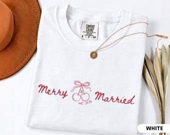May include: A white t-shirt with a red graphic print featuring the text Merry & Married, a pair of cherries tied with a bow, and the text est. 2026. The shirt has a Comfort Colors brand label at the neck with size S-P-CH and a gold-toned necklace.
