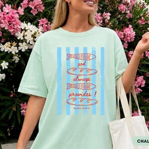 Woman wearing oversized Comfort Colors Christians shirt featuring God Always Provides graphic with fish illustrations and Matthew 14:13-21 Bible verse in vintage aesthetic style.