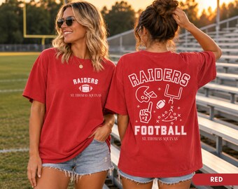 May include: A red short-sleeve t-shirt featuring white graphics. The front displays "RAIDERS" above a football and "ST. THOMAS AQUINAS". The back shows "RAIDERS FOOTBALL" with a helmet, goalpost, football, foam finger, and play diagrams.