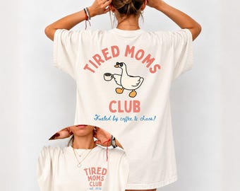 May include: An off-white t-shirt featuring a graphic of a goose wearing sunglasses and holding a coffee mug. The back text reads TIRED MOMS CLUB and Fueled by coffee & chaos! The front chest area displays TIRED MOMS CLUB and est. 2024.