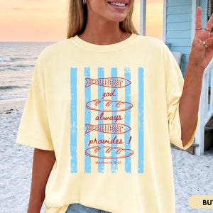 Woman wearing oversized Comfort Colors Christians shirt featuring God Always Provides graphic with fish illustrations and Matthew 14:13-21 Bible verse in vintage aesthetic style.